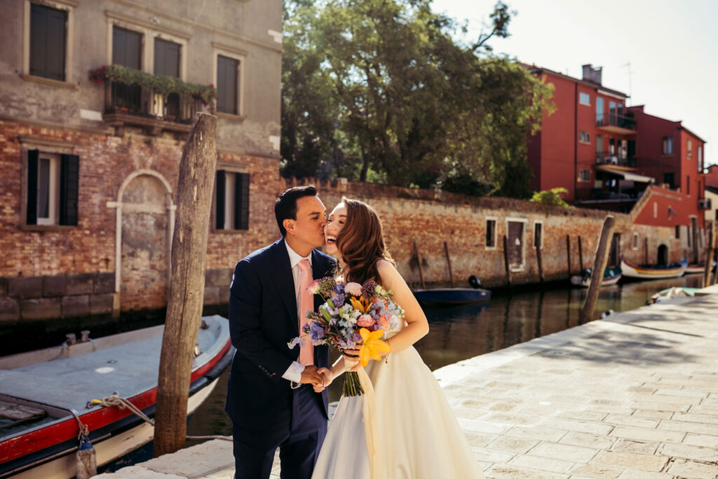 The groom kisses the bride, beautifully captured in this timeless portrait within the enchanting backdrop of the Venetian canals