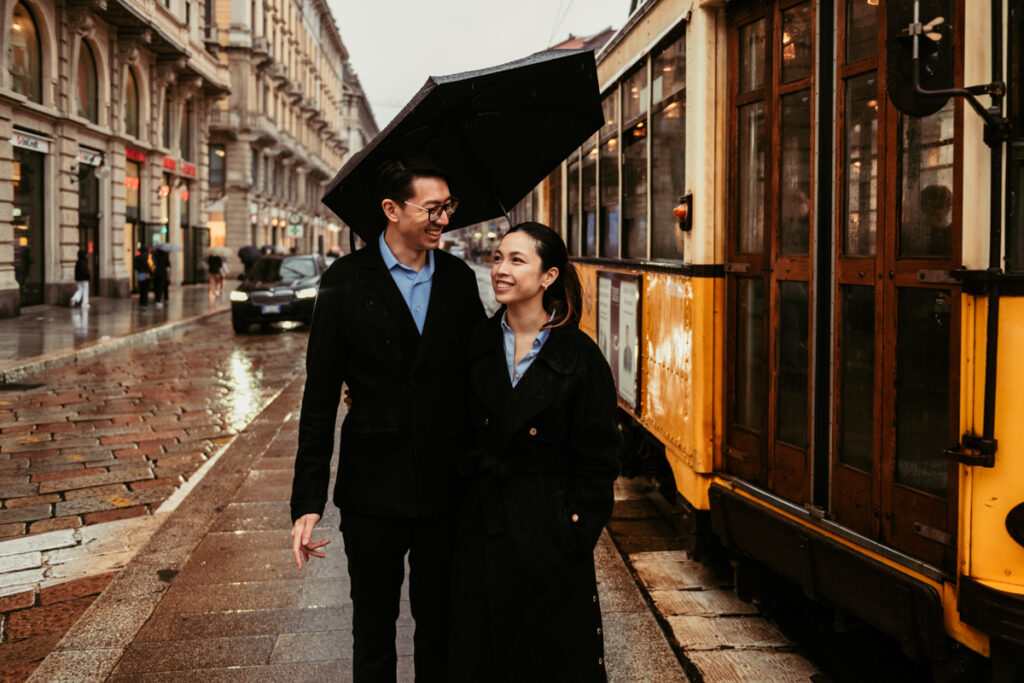 Couple laughing under an umbrella through Milan streets as a vintage tram passes by