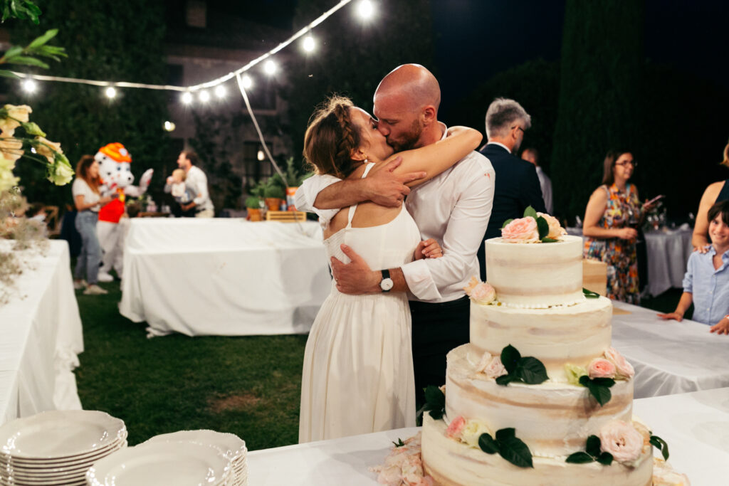 Bride and groom kissing in front of the wedding cake adorned with fresh flowers at a Langhe vineyard wedding