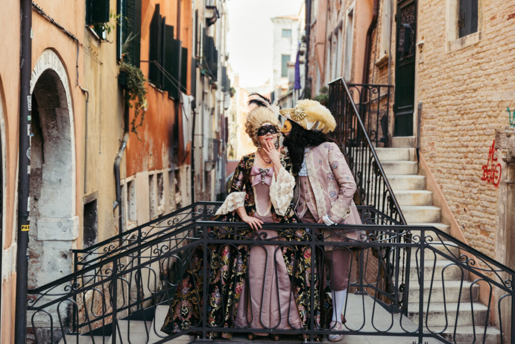 Couple in 18th-century costumes on a Venetian bridge, as a man kisses a woman on the cheek in a romantic moment.