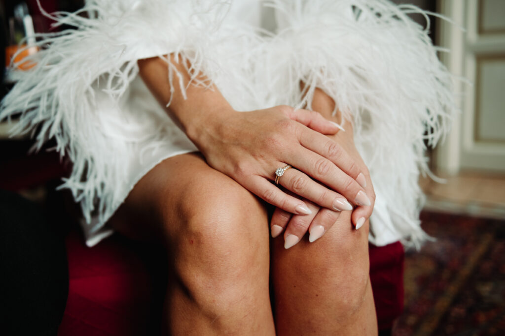 Close-up of the bride’s hands resting on her lap, showing her engagement ring and feathered robe details.