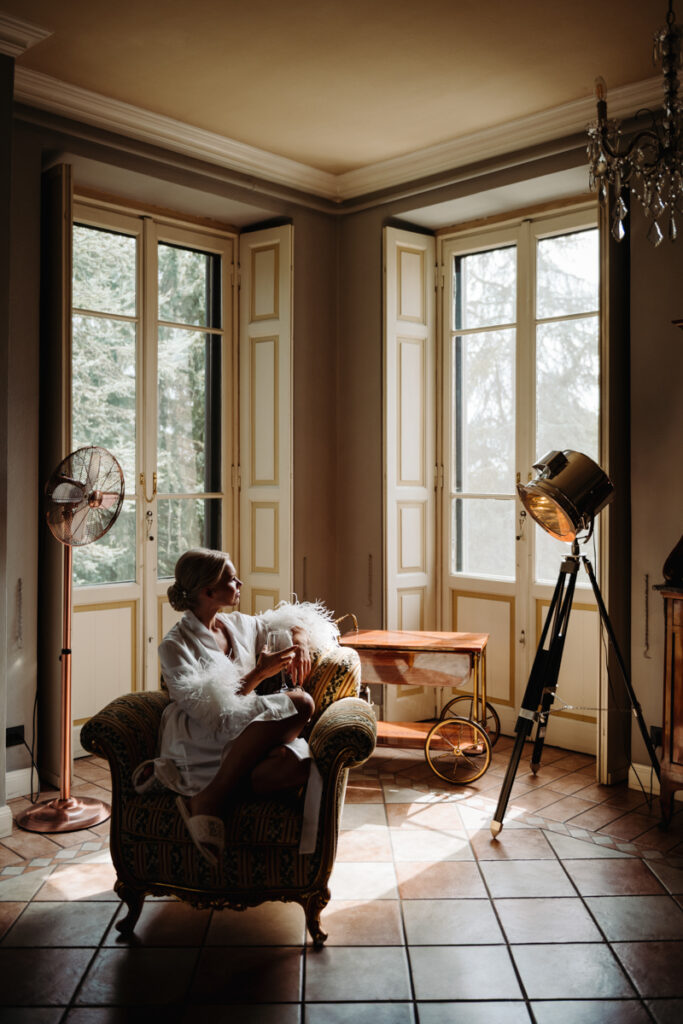 Bride relaxing in a cozy room with large windows, enjoying a quiet moment before the ceremony.