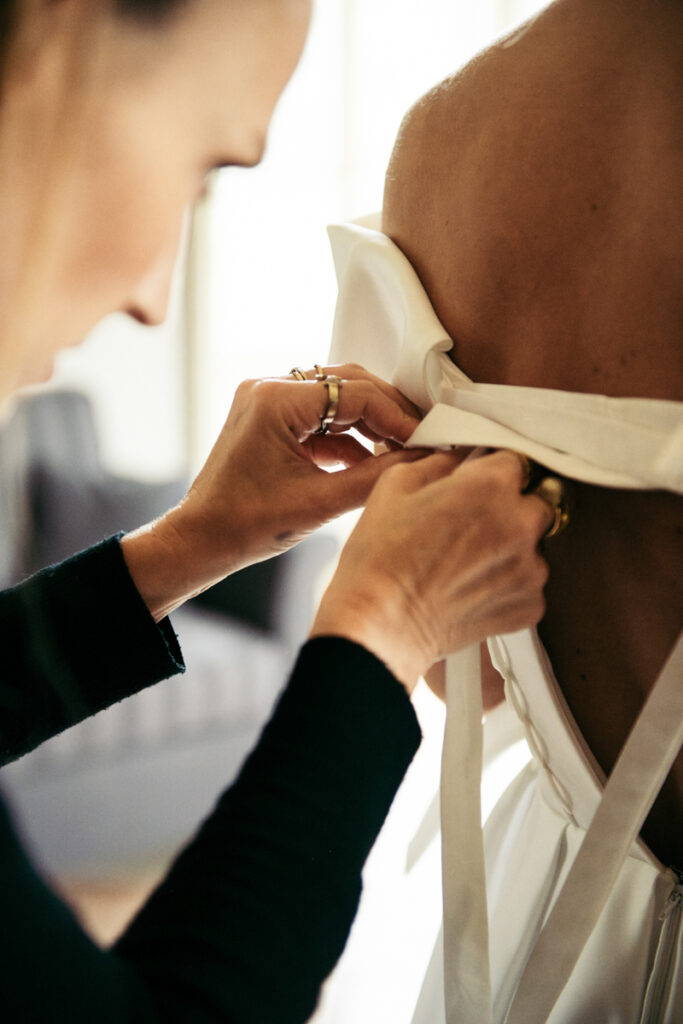 Close-up of hands carefully tying the ribbon on the back of the bride’s dress during the final touches.