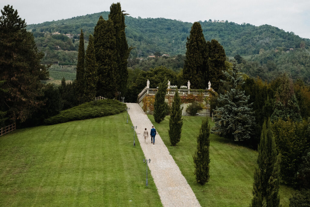 Aerial view of a tree-lined path leading to an Italian villa, with the bride and groom walking hand in hand.