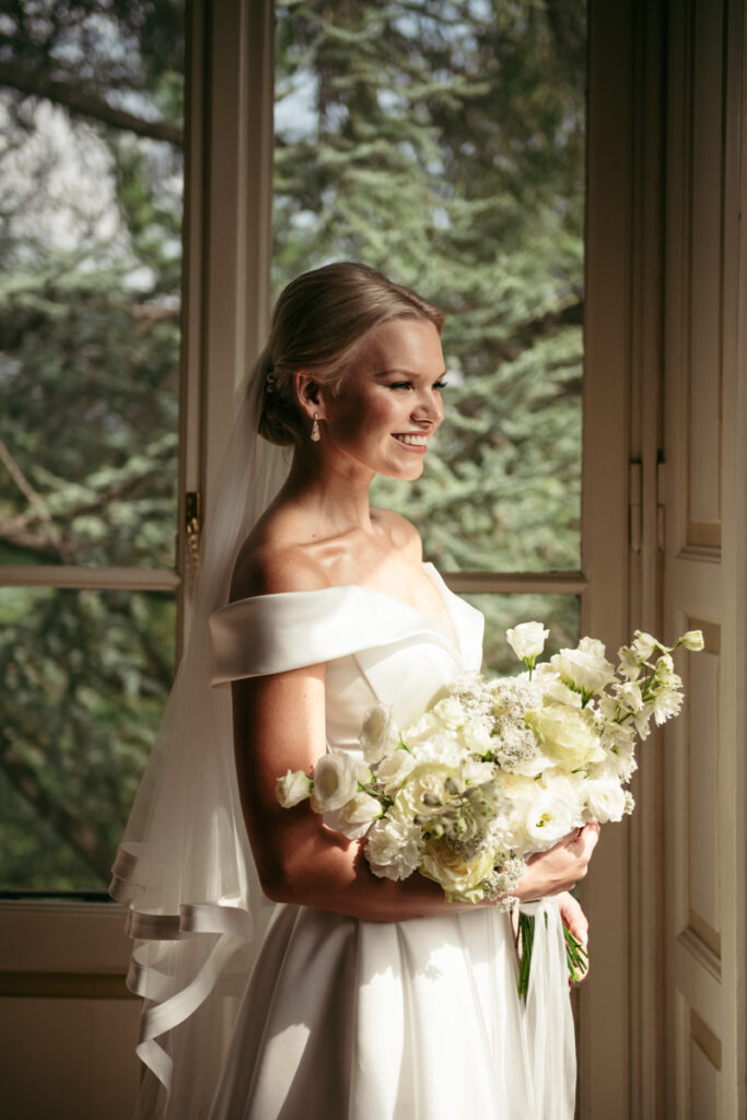 Bride looking thoughtfully out of the window, her white bouquet held gently in her hands.