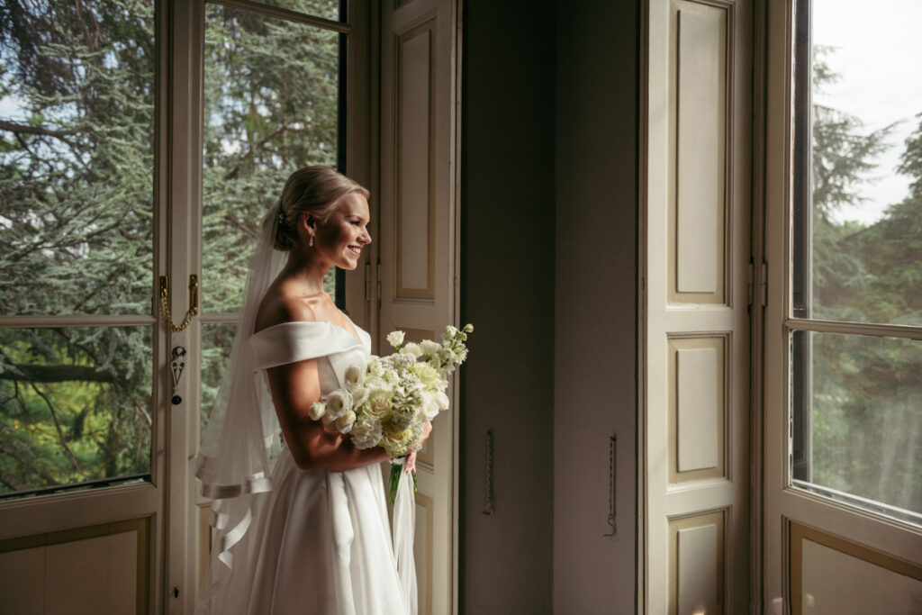 Bride standing by a large window, holding her bouquet and gazing outside with a serene expression.