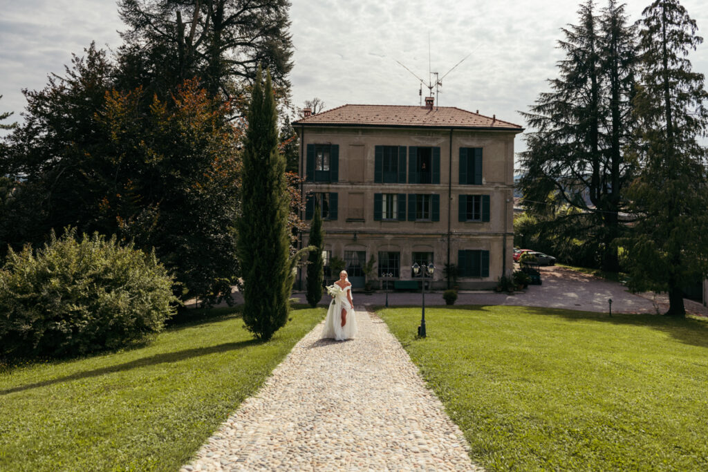 Bride walking down a gravel path toward the villa, framed by tall cypress trees and elegant gardens.