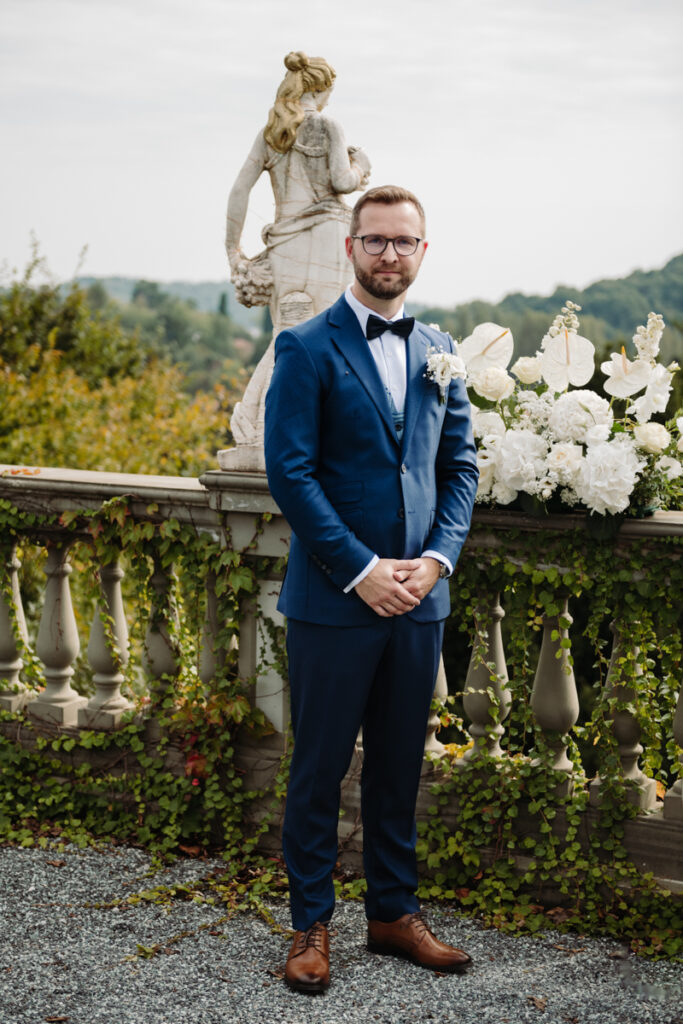 Groom standing confidently, smiling softly with a backdrop of white flowers and greenery.