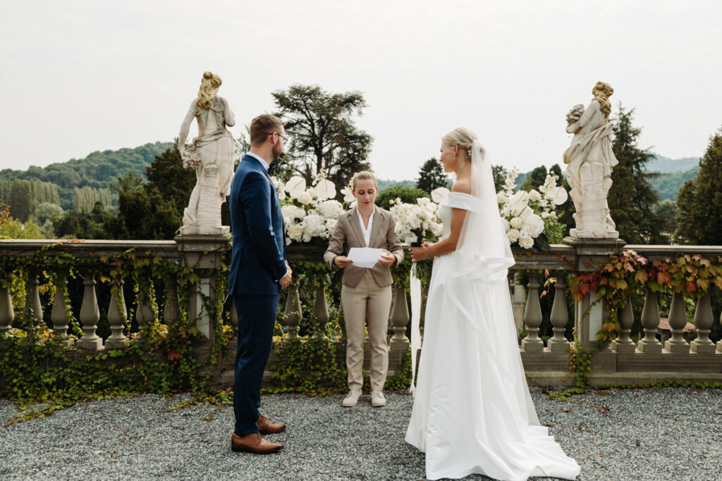 The couple exchanging vows in front of lush white floral decorations and statues.