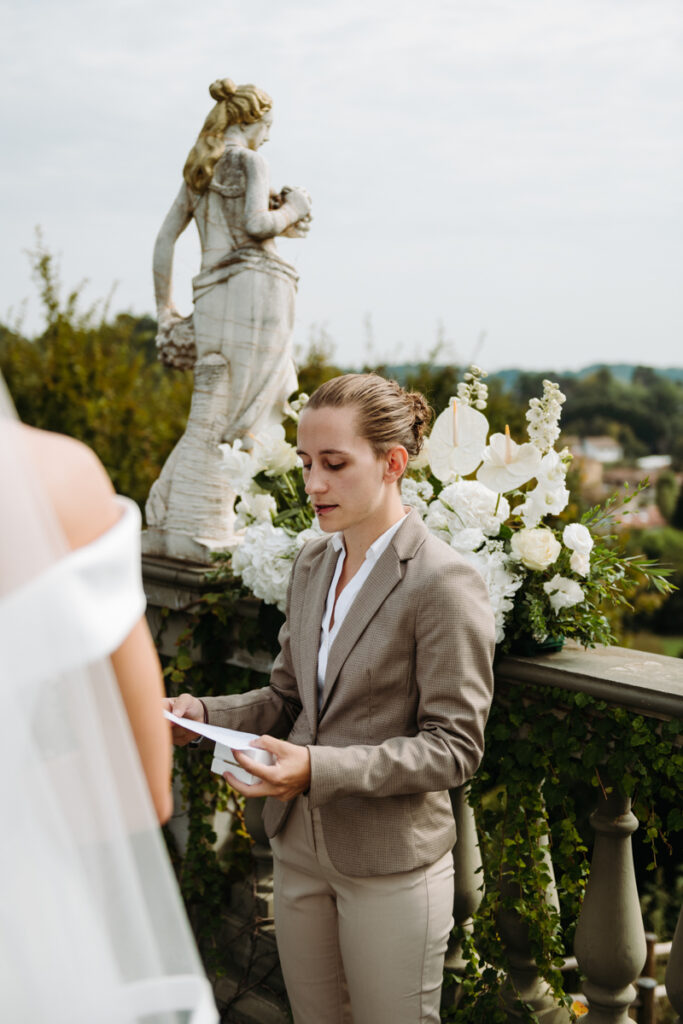 The celebrant reading vows during the symbolic ceremony, surrounded by white flowers and statues.