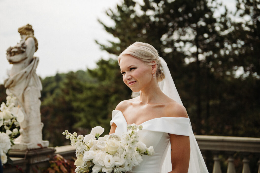 Bride smiling softly while holding a white floral bouquet, with greenery in the background.