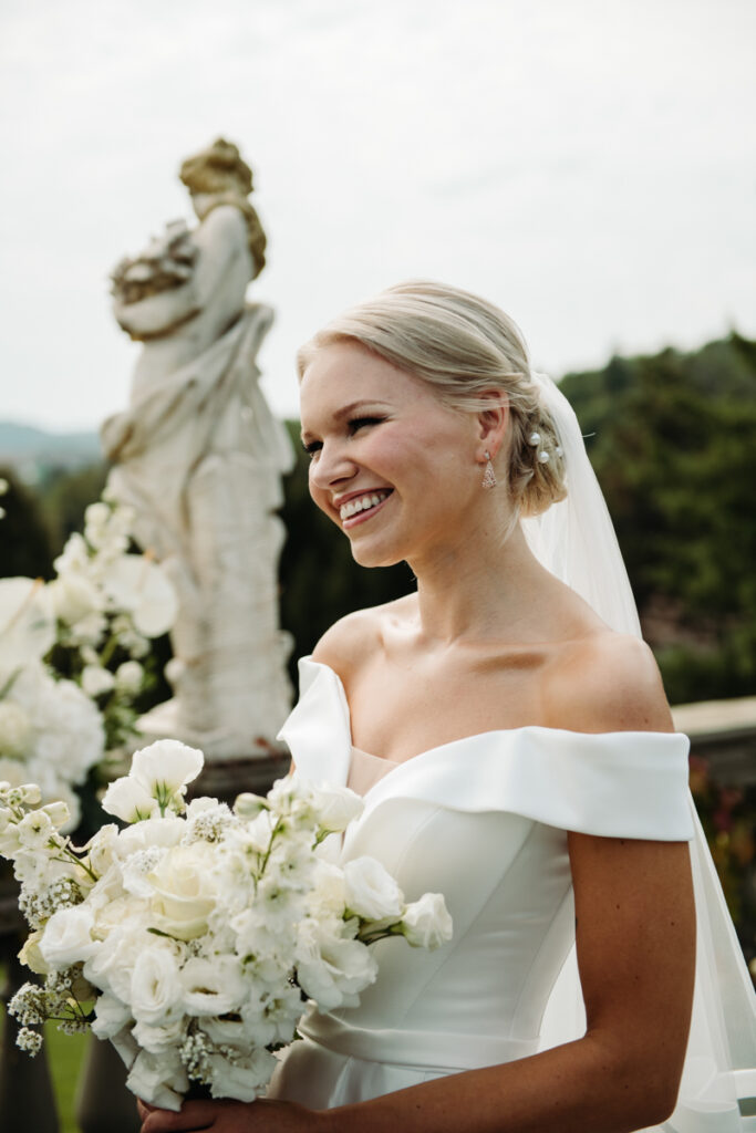 Bride smiling joyfully, her bouquet in hand, with classical statues and white flowers in the background.