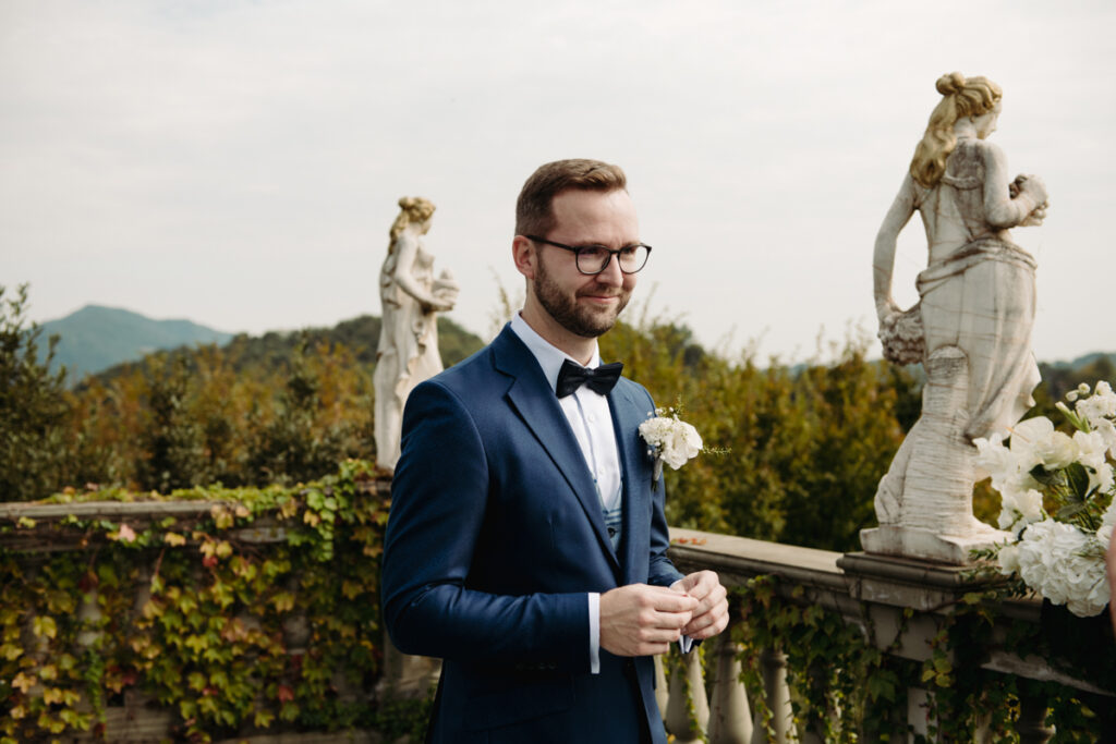 Groom standing confidently with a soft smile, holding his boutonnière against a scenic Italian backdrop.