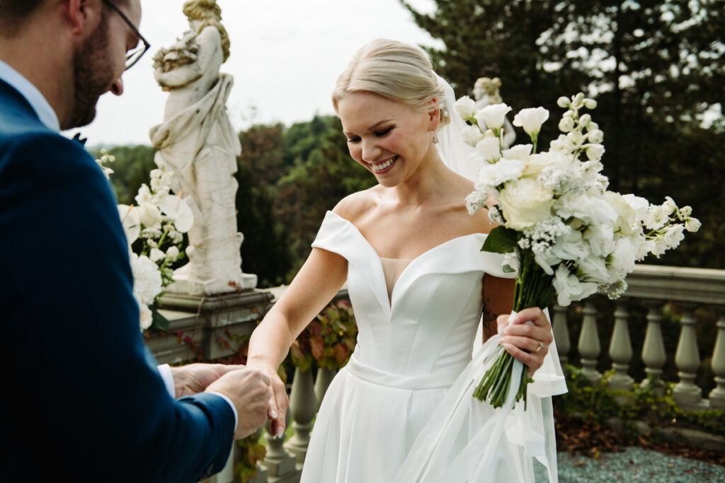 Bride smiling warmly while holding her bouquet, while the groom is putting on her ring