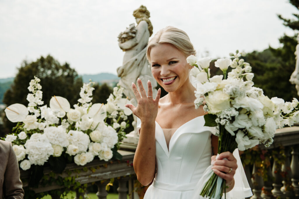 The bride smiling brightly and showing her ring, surrounded by beautiful white floral arrangements.