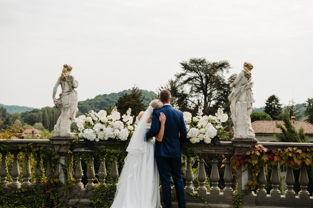 Bride and groom embracing on a terrace decorated with white flowers, surrounded by classical statues