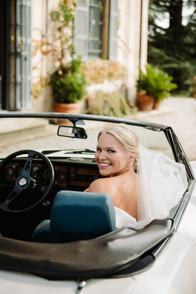 Bride smiling while sitting in the driver’s seat of a vintage convertible