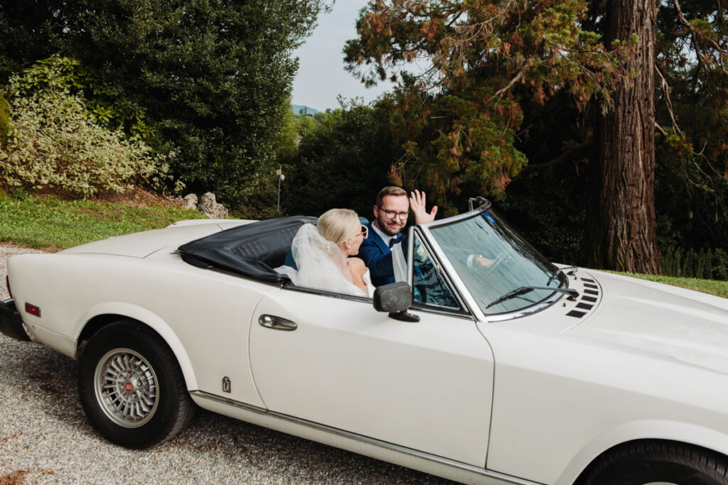 Bride and groom sitting in a vintage white convertible, smiling at each other