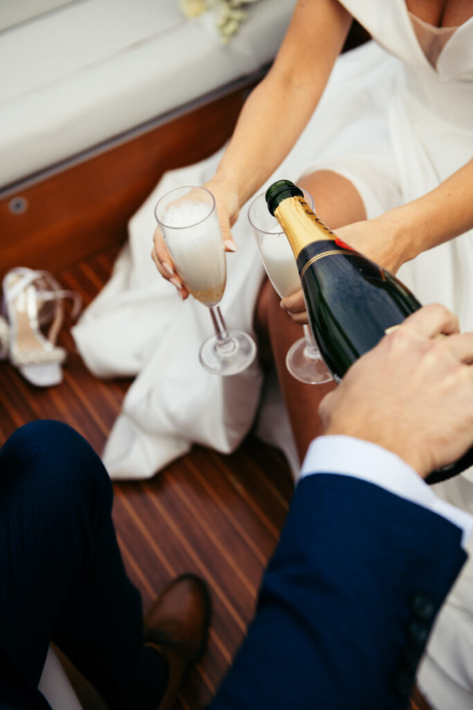 Close-up of hands opening a bottle of champagne, with bridal shoes on the deck