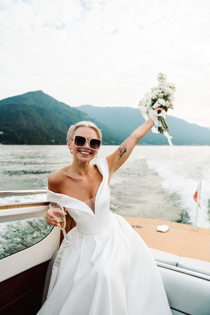 Bride raising her bouquet and a champagne glass, celebrating on a boat in Lake Como