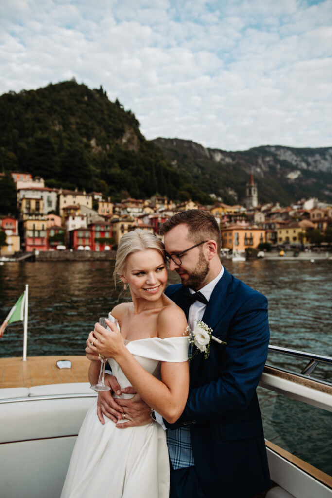 Groom embracing the bride while standing on a boat with the town of Varenna in the background