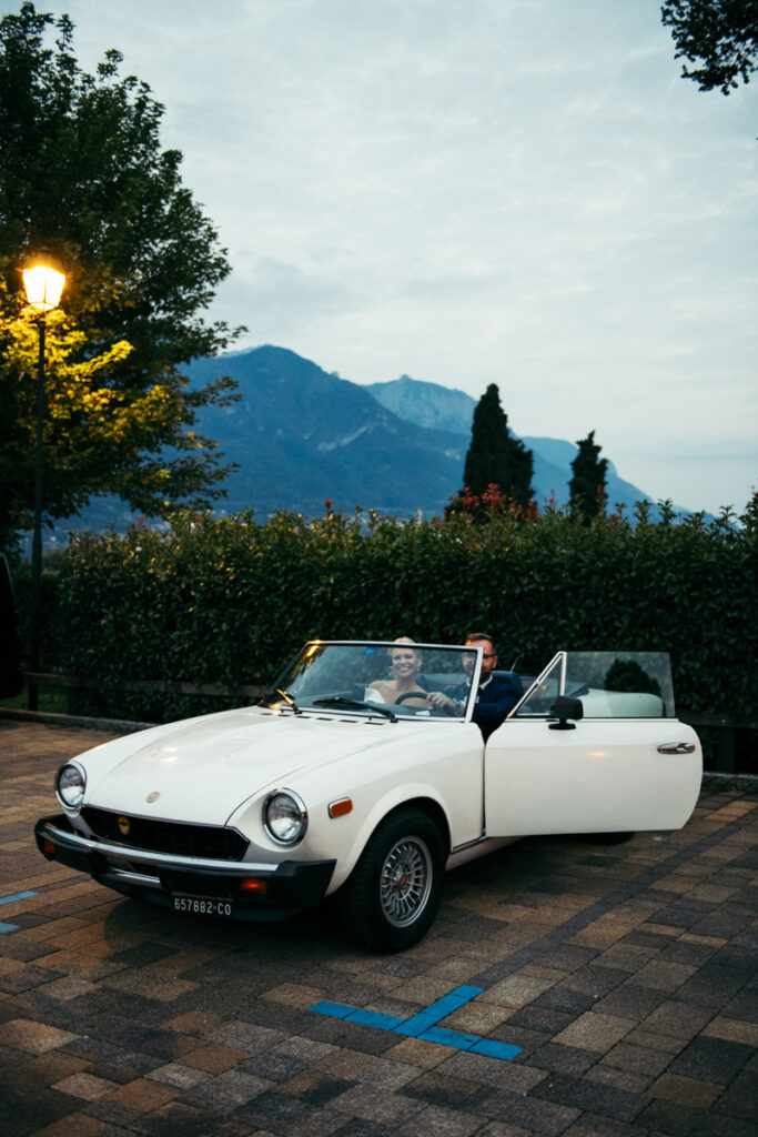 Bride and groom driving a vintage white convertible with scenic mountains in the background.