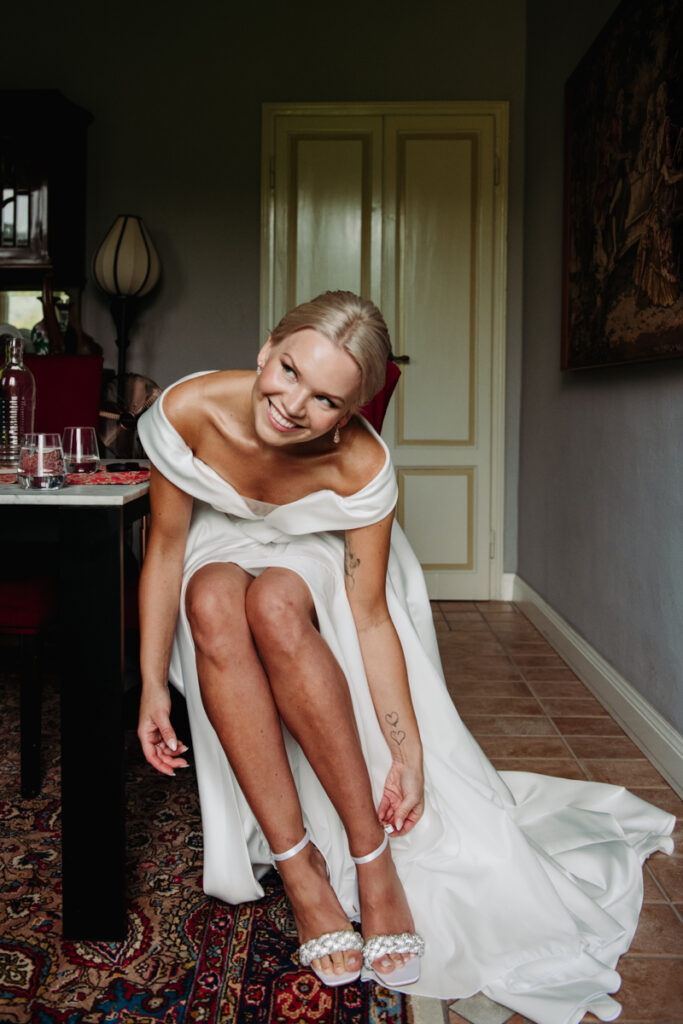 Bride sitting down and laughing while adjusting her shoes indoors