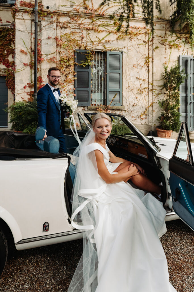 Bride smiling while stepping out of a vintage car, with the groom standing nearby