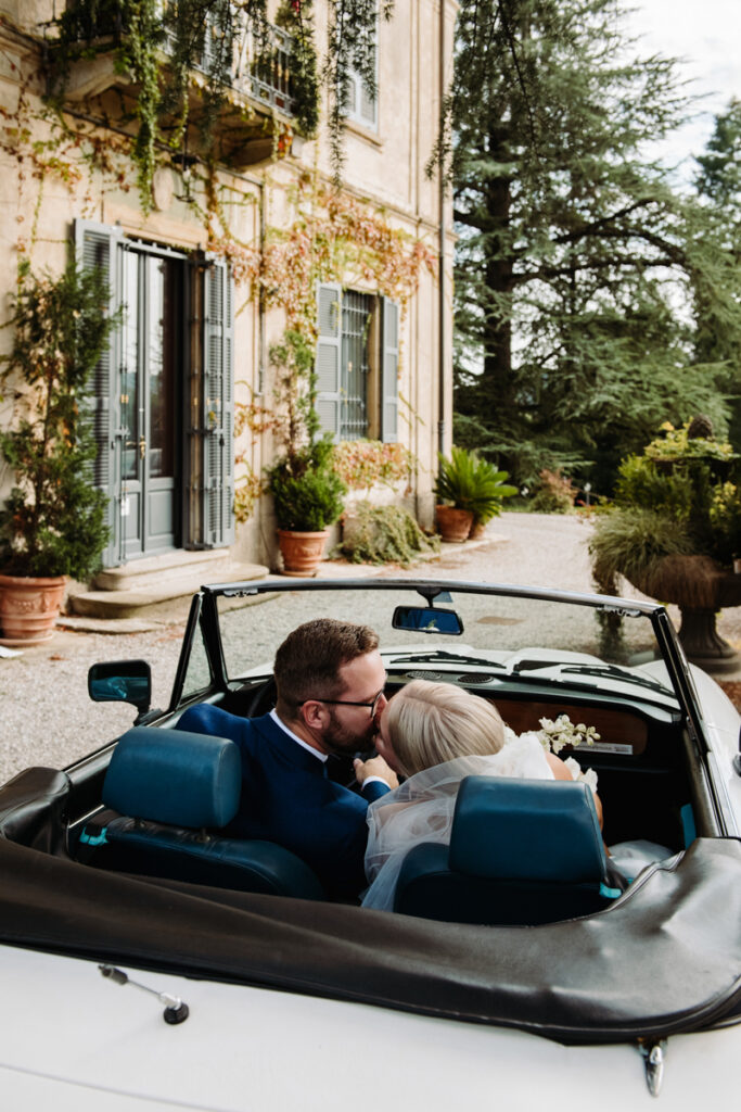 Bride and groom sharing a kiss in a vintage convertible parked outside an Italian villa