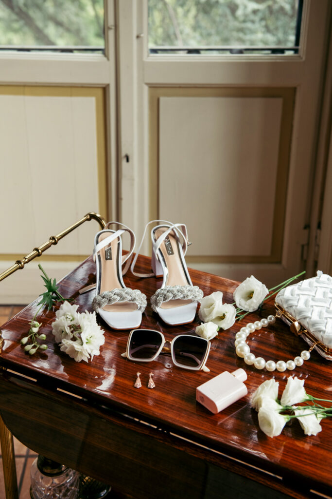  A beautifully arranged flat lay of bridal accessories on a polished wooden table, featuring white braided-strap heels, a pearl necklace, a white quilted handbag, sunglasses, delicate earrings, a perfume bottle, and fresh white flowers. A refined and elegant detail shot for an intimate elopement