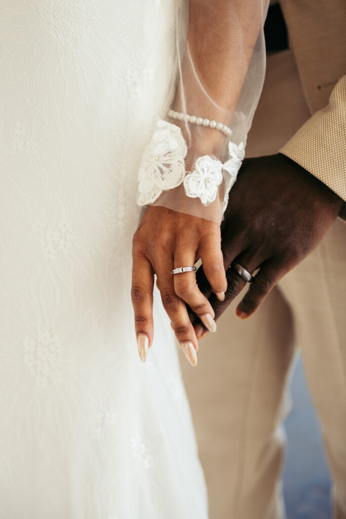 Bride's hand adorned with a lace glove and wedding ring, holding her groom’s hand.