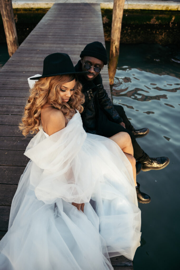 Bride in a black hat and groom in casual outfit sitting on a wooden dock in Venice, with a dreamy look.