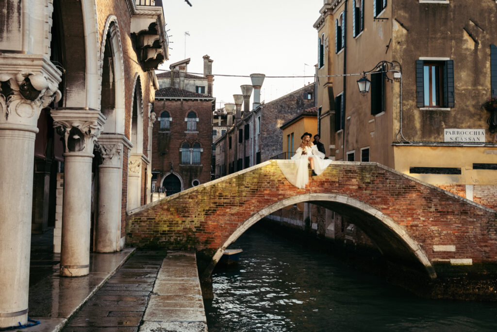 Groom and bride on a historic Venice bridge, with pastel-colored buildings behind them.