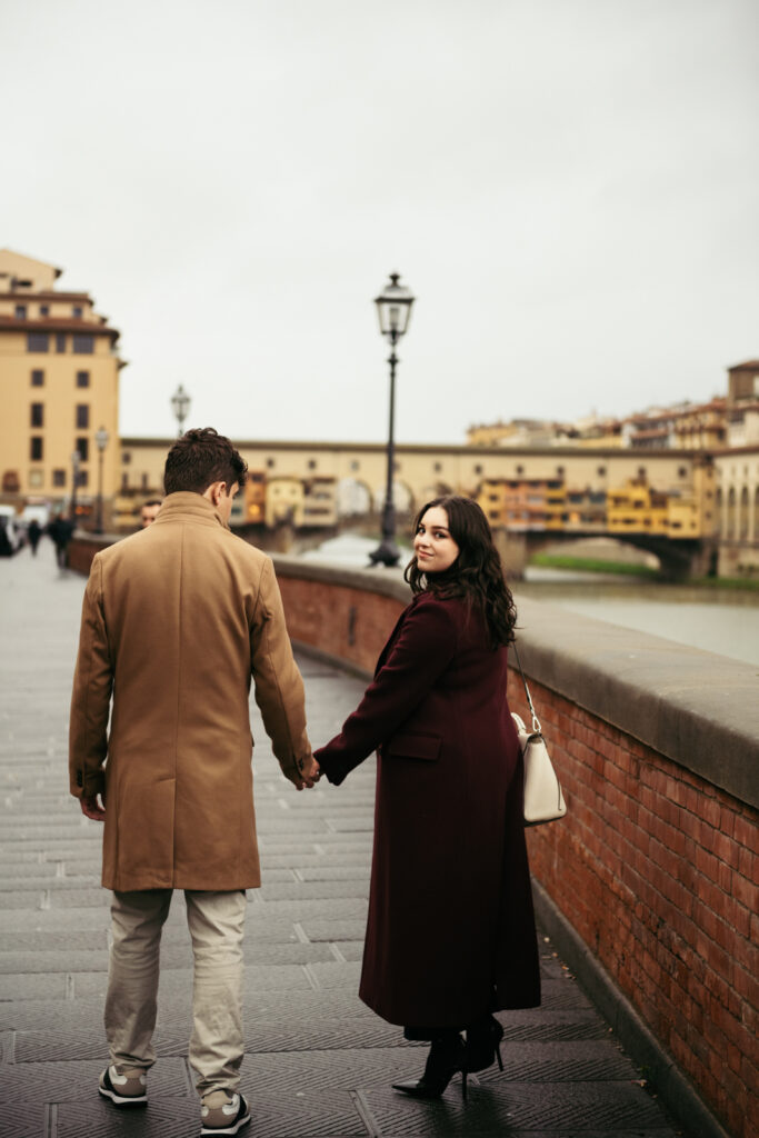 A couple walking along the Arno River in Florence, with the woman glancing at the camera while holding hands with her partner, a candid and romantic moment.