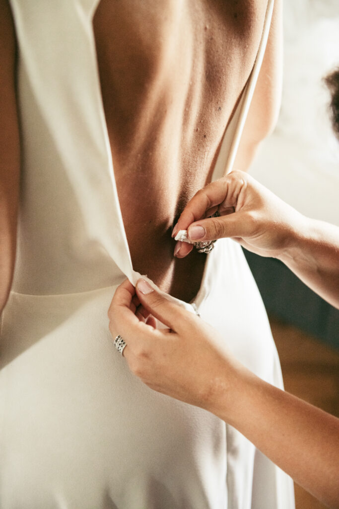 Bridesmaid zipping up the bride’s elegant dress during the getting ready