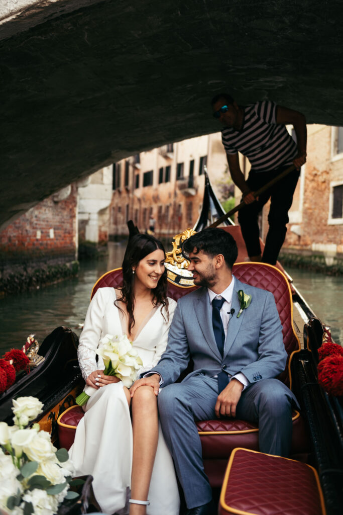 Bride and groom smiling during symbolic wedding on a gondola under a bridge in Venice