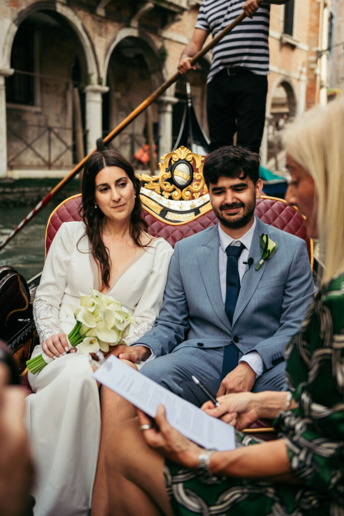 Bride and groom seated on a gondola during symbolic wedding in Venice, listening to the officiant
