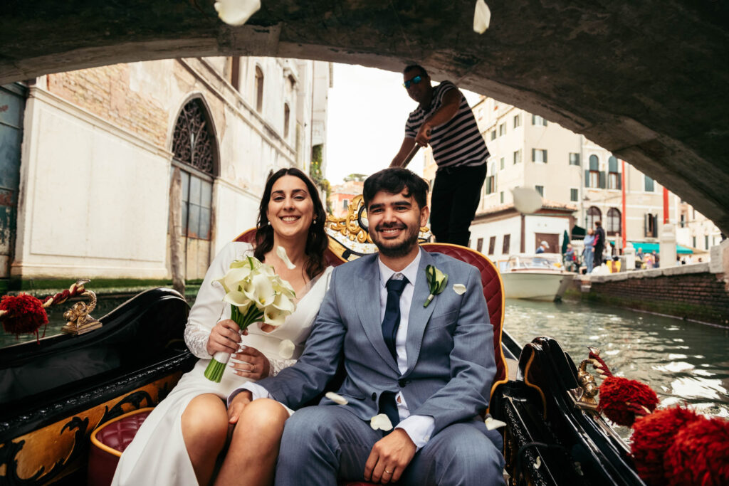 Bride and groom seated in a gondola under a stone bridge during their symbolic wedding in Venice